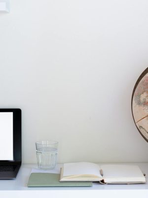 Creative composition of computer with empty screen placed near notebook with white papers and globe with glass of water in between